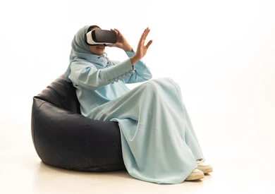 المتعة والتسلية بالمشاهدة عبر نظارة الواقع الافتراضي,بورترية لإمراة عربية خليجية سعودية محجبة ترتدي عباءة تجلس على كرسي بين باج  تضع نظارات ثلاثية الأبعاد تقوم برفع كف يدها للاعلى بتعابير الاندماج والتوقف عن فعل امر ما, خلفية بيضاء