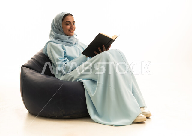 The culture of self-learning, gestures of integration and focus, spending time learning and educating oneself, enjoying reading books, a portrait of a Saudi Arabian Gulf woman wearing a hijab and an abaya sitting on a chair between bags holding a book in her hands, white background.