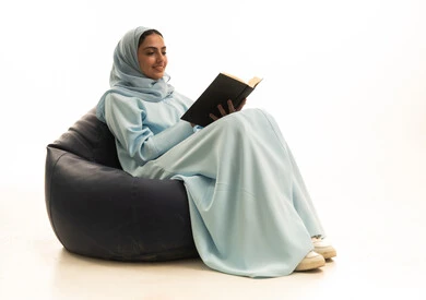 ثقافة التعلم الذاتي، ايماءات الاندماج و التركيز، قضاء الوقت بالتعلم والتثقف، الاستمتاع بقراءة الكتب ، بورترية لإمراة عربية خليجية سعودية محجبة ترتدي عباءة تجلس على كرسي بين باج تحمل بيديها كتاب، خلفية بيضاء