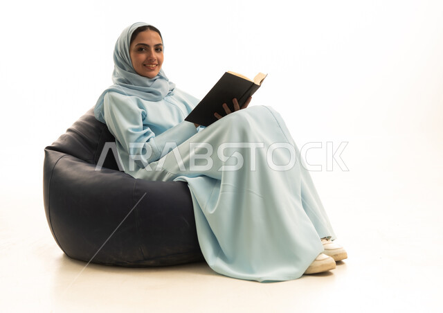 النظر للكاميرا بتعابير السرور، ثقافة التعلم الذاتي، قضاء الوقت بالتعلم والتثقف، الاستمتاع بقراءة الكتب ، بورترية لإمراة عربية خليجية سعودية محجبة ترتدي عباءة تجلس على كرسي بين باج تحمل بيديها كتاب، خلفية بيضاء