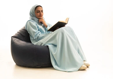 Gestures of integration and focus, the culture of self-learning, spending time learning and educating oneself, enjoying reading books, a portrait of a Saudi Arabian Gulf woman wearing a hijab and an abaya sitting on a chair between bags holding a book in her hands, white background.