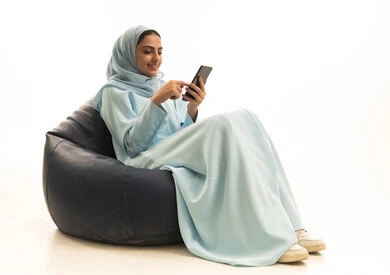 تصفح مواقع التواصل الاجتماعي عبر الانترنت، الدردشة مع الاهل و الاصدقاء، بورتريه لامراة عربية خليجية سعودية ترتدي الحجاب والعباءة تجلس على كرسي بين باج تحمل بيدها الهاتف المحمول ، استخدام الأجهزة التقنية الحديثة، خلفية بيضاء