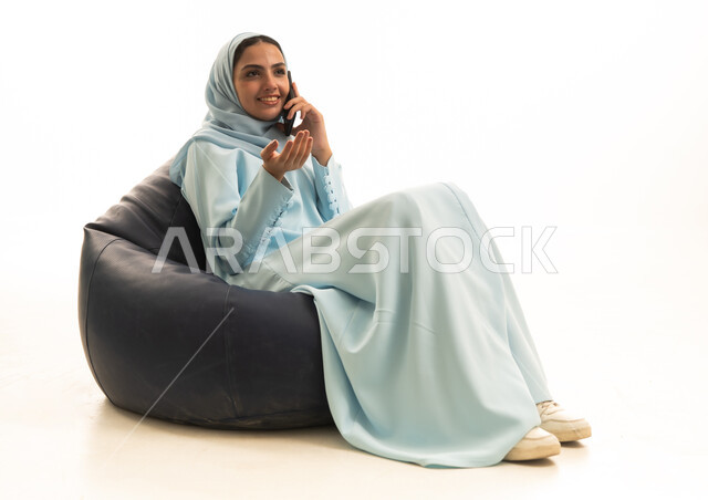 اجراء مكالمة هاتفية عبر الجوال، الدردشة مع الاهل و الاصدقاء، بورتريه لامراة عربية خليجية سعودية ترتدي الحجاب والعباءة تجلس على كرسي بين باج تحمل بيدها الهاتف المحمول ، استخدام الأجهزة التقنية الحديثة، خلفية بيضاء