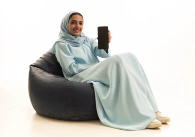 عرض شاشة سوداء فارغة ، بورتريه لامراة عربية خليجية سعودية ترتدي الحجاب والعباء تجلس على كرسي بين باج تحمل بيدها الهاتف المحمول ، استخدام الأجهزة التقنية الحديثة، خلفية بيضاء