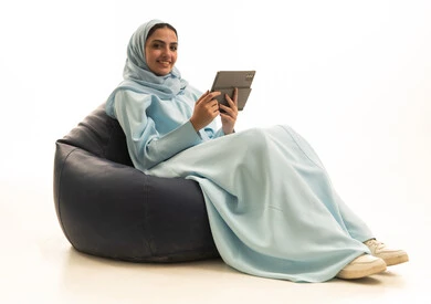 دمج التقنيات الحديثة والتكنولوجيا في الحياة العملية، استخدام التقنية الحديثه المتطورة في الحياة العملية عبر الجهاز اللوحي، تعابير وايماءات الاندماج، بورتريه لامراة عربية خليجية سعودية ترتدي الحجاب والعباء تجلس على كرسي بين باج  تقوم بالعمل على جهازالتابلت، خلفية بيضاء