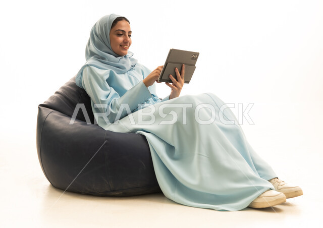 استخدام التقنية الحديثه المتطورة في الحياة العملية عبر الجهاز اللوحي، تعابير وايماءات الاندماج، بورتريه لامراة عربية خليجية سعودية ترتدي الحجاب والعباء تجلس على كرسي بين باج  تقوم بالعمل على جهازالتابلت، خلفية بيضاء