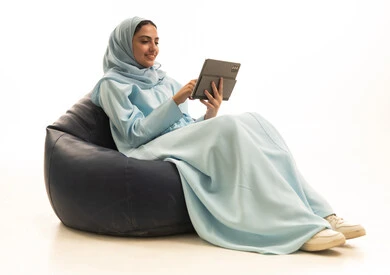 The use of advanced modern technology in professional life through a tablet, expressions and gestures of integration, a portrait of a Gulf Arab Saudi woman wearing a hijab and abaya sitting on a chair between bags working on a tablet, white background.