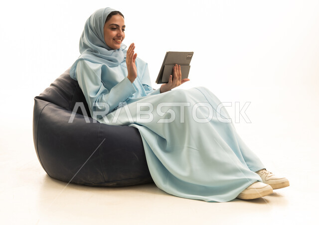 اجراء مكالمة فيديو عبر الانترنت، رفع اليد بتعابير السلام و التحية، استخدام التقنية الحديثه المتطورة في الحياة العملية عبر الجهاز اللوحي، تعابير وايماءات الاندماج، بورتريه لامراة عربية خليجية سعودية ترتدي الحجاب والعباء تجلس على كرسي بين باج  تقوم بالعمل على جهاز التابلت، خلفية بيضاء