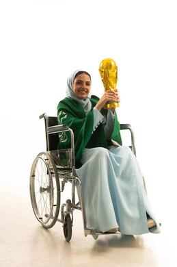 اليوم الوطني السعودي 23 سبتمبر، مشاركة النساء السعوديات في الأنشطة الرياضية والمباريات، تشجيع الأنديه والمنتخبات، بورتريه لشابة عربية سعودية خليجية ترتدي العباءة و علم السعودية تجلس على كرسي متحرك تحمل الكأس ترفع يدها بإيماءات الحماس لفوز وانتصار الفريق المفضل، خلفية بيضاء