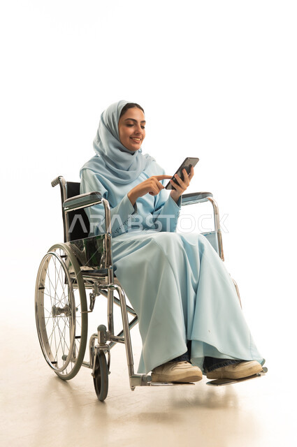إيماءات السعادة والسرور، الدردشة مع الأهل والأصدقاء، استخدام الاجهزة التقنية الحديثة، بورتريه لامراه عربية خليجية سعودية من ذوي الاحتياجات الخاصة محجبة ترتدي العباءة تجلس على كرسي متحرك تحمل بيدها الهاتف المحمول، تصفح وسائل التواصل الاجتماعي، خلفية بيضاء