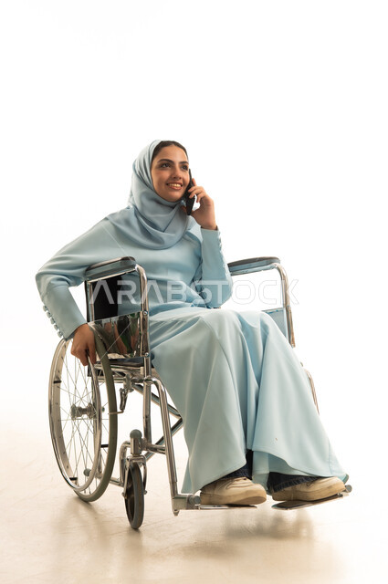 استخدام الأجهزة التكنولوجية الحديثة والمتطورة، بورتريه لامراه عربية خليجية سعودية من ذوي الاحتياجات الخاصة محجبة ترتدي العباءة تجلس على كرسي متحرك تقوم بالدردشة والتحدث على الهاتف المحمول، النظر إلى شيء ما أثناء إجراء مكالمة هاتفية عبر الجوال، خلفية بيضاء