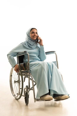 استخدام الأجهزة التكنولوجية الحديثة والمتطورة، بورتريه لامراه عربية خليجية سعودية من ذوي الاحتياجات الخاصة محجبة ترتدي العباءة تجلس على كرسي متحرك تقوم بالدردشة والتحدث على الهاتف المحمول، النظر إلى شيء ما أثناء إجراء مكالمة هاتفية عبر الجوال، خلفية بيضاء
