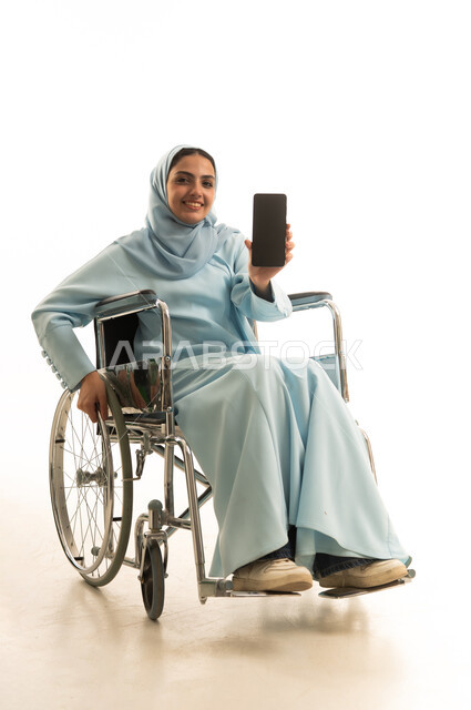 عرض تطبيقات الهاتف المحمول، التسويق الالكتروني باستخدام التقنية والتكنولوجيا، بورتريه لامراه عربية خليجية سعودية من ذوي الاحتياجات الخاصة محجبة ترتدي العباءة تجلس على كرسي متحرك تحمل بيدها جوال بشاشة سوداء فارغة، خلفية بيضاء
