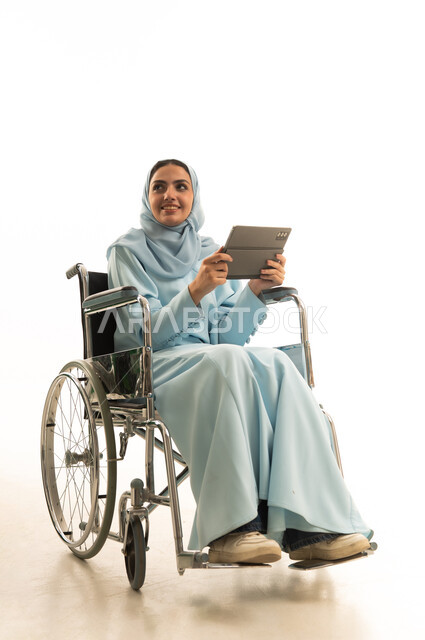 التعلم الإلكتروني من خلال استخدام التقنيات الحديثة، إجراء مكالمات الفيديو والبثوث المباشرة، متابعة الأفلام والمسلسلات المفضلة عبر الإنترنت، النقر على شاشة الجهاز اللوحي، بورتريه لامراه عربية خليجية سعودية من ذوي الاحتياجات الخاصة محجبة ترتدي العباءة تجلس على كرسي متحرك تحمل جهاز التابلت المحمول، خلفية بيضاء