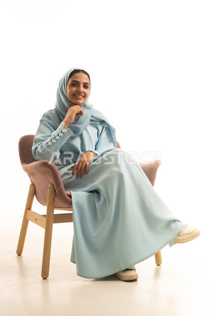 Trying to find a solution to something, contemplation, focus, and planning for the future, gestures of confusion and anxiety, staring blankly at something, a portrait of a Saudi Arabian Gulf woman wearing a hijab and an abaya sitting on a chair with her hand on her chin, looking thoughtfully and reflectively, with a white background.