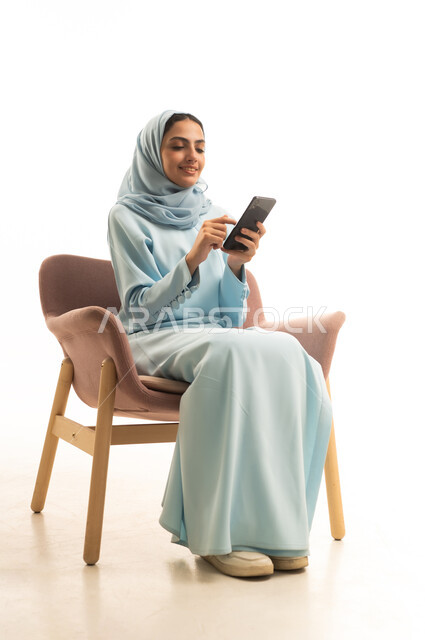 Chatting with family and friends, gestures of focus and engagement, using a modern and advanced technical device, a portrait of a Saudi Arabian Gulf woman wearing a hijab and an abaya sitting on a chair holding a mobile phone, browsing social media on her phone, white background.
