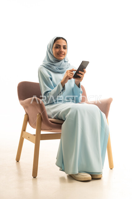 إيماءات السعادة و السرور، الدردشة مع الأهل والأصدقاء، استخدام جهاز تقني حديث ومتطور، بورتريه لامرأة عربية خليجية سعودية محجبة ترتدي العباءة تجلس على كرسي تحمل بيدها الهاتف المحمول، تصفح وسائل التواصل الاجتماعي عبر الجوال، خلفية بيضاء