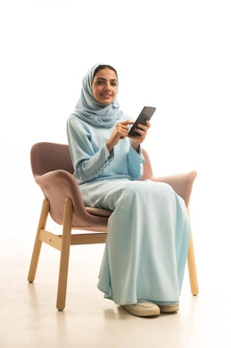 Gestures of happiness and joy, chatting with family and friends, using a modern and advanced technological device, a portrait of a Saudi Arabian Gulf woman wearing a hijab and an abaya sitting on a chair holding a mobile phone, browsing social media on her phone, white background.