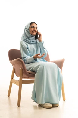 استخدام الأجهزة التقنية الحديثة في الحياة اليومية,التواصل مع العائلة والأصدقاء, مشاركة الأخبار والأحاديث الجيدة, بورتريه لامرأة عربية خليجية سعودية محجبة ترتدي العباءة تجلس على كرسي تمسك بيدها الهاتف المحمول،إجراء اتصال هاتفي, خلفية بيضاء