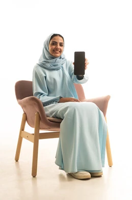 عرض شاشة سوداء فارغة ، بورترية لامراه عربيه خليجيه سعودية ترتدي العباءة و الحجاب تجلي على كرسي تحمل بيدها الهاتف المحمول ، استخدام الأجهزة التقنية الحديثة، خلفية بيضاء