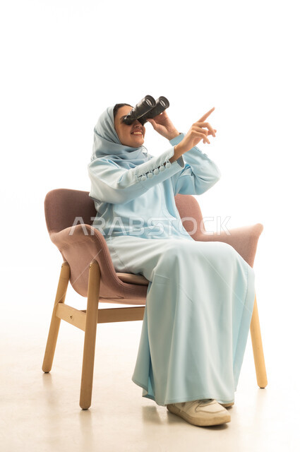 الاشارة باصبع السبابة الى مكان ما، رؤية الأشياء البعيدة، التخطيط المستقبلي للأهداف والطموحات، التفكير والتطلع إلى المستقبل، حب الاستكشاف والمغامرة، بورتريه لشابة عربية خليجية سعودية محجبة ترتدي العباءة تجلس على كرسي تمسك منظار ثنائي العينين، خلفية بيضاء
