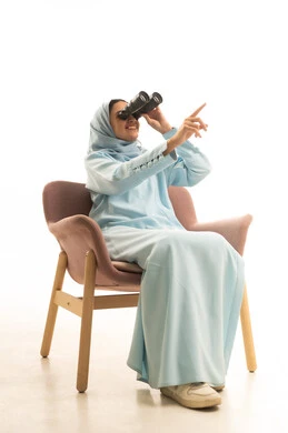Pointing with the index finger to a certain place, seeing distant things, future planning for goals and ambitions, thinking and looking forward to the future, love for exploration and adventure, a portrait of a young Arab Gulf Saudi woman wearing a hijab and an abaya sitting on a chair holding binoculars, white background.
