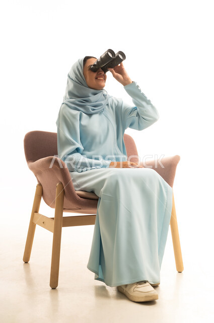رؤية الأشياء البعيدة، التخطيط المستقبلي للأهداف والطموحات، التفكير والتطلع إلى المستقبل، حب الاستكشاف والمغامرة، بورتريه لشابة عربية خليجية سعودية محجبة ترتدي العباءة تجلس على كرسي تمسك منظار ثنائي العينين، خلفية بيضاء