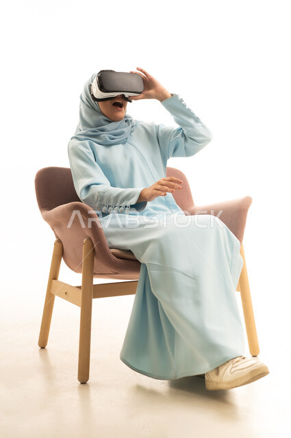 التفاعل مع فيديو ثلاثي الأبعاد، استخدام التكنولوجيا الحديثة، محاكاة العالم الخيالي، بورتريه لإمرأة عربية خليجية سعودية محجبة ترتدي العباءة  تجلس على كرسي وتضع نظارة الواقع الافتراضي ترفع يديها بايماءات الدهشة والاستغراب، خلفيه بيضاء