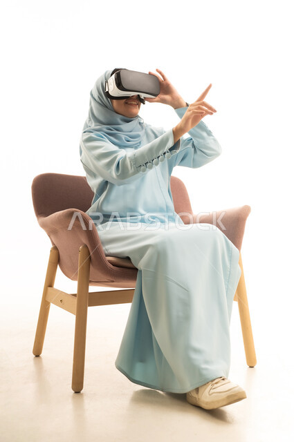 The enjoyment of watching through virtual reality glasses, pointing to a specific place, a portrait of a Gulf Arab Saudi woman wearing an abaya sitting on a chair wearing VR glasses, pointing with her index fingers sideways with expressions of this place here, simulating the imaginary world, white background.
