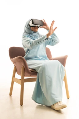 Raising hands with gestures of immersion and enjoyment, simulating a fantasy world, a portrait of a Saudi Arabian Gulf woman wearing a hijab and an abaya sitting on a chair, wearing 3D glasses and moving and interacting with it, watching movies using virtual reality technologies, white background.