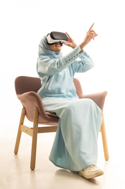 Pointing to a specific place, the enjoyment of viewing through virtual reality glasses, a portrait of a Saudi Gulf Arab woman wearing an abaya sitting on a chair wearing VR glasses, pointing with her index fingers sideways with expressions of this place here, simulating the imaginary world, white background.