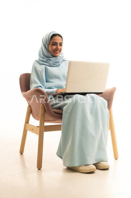 استخدام جهاز تقني حديث ومتطور، ادارة العمل عن بعد، بورتريه لامرأة عربية خليجية سعودية محجبة ترتدي العباءة تجلس على كرسي تعمل على جهاز الحاسوب المحمول, إنجاز المهام المطلوبة عبر اللابتوب، خلفية بيضاء
