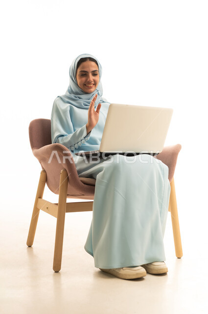 رفع كف اليد بتعابير التحية و السلام ، اجراء مكالمة فيديو عبر الانترنت، استخدام جهاز تقني حديث ومتطور، ادارة العمل عن بعد، بورتريه لامرأة عربية خليجية سعودية محجبة ترتدي العباءة تجلس على كرسي تعمل على جهاز الحاسوب المحمول, إنجاز المهام المطلوبة عبر اللابتوب، خلفية بيضاء