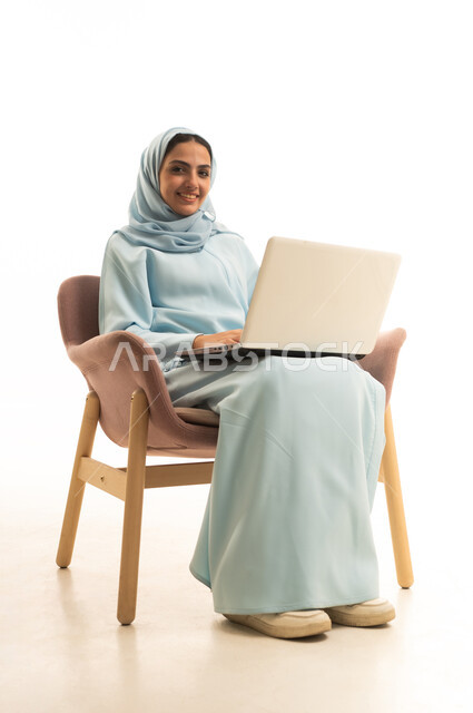 ادارة العمل عن بعد، استخدام جهاز تقني حديث ومتطور، بورتريه لامرأة عربية خليجية سعودية محجبة ترتدي العباءة تجلس على كرسي تعمل على جهاز الحاسوب المحمول, إنجاز المهام المطلوبة عبر اللابتوب، خلفية بيضاء