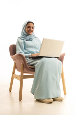 ادارة العمل عن بعد، استخدام جهاز تقني حديث ومتطور، بورتريه لامرأة عربية خليجية سعودية محجبة ترتدي العباءة تجلس على كرسي تعمل على جهاز الحاسوب المحمول, إنجاز المهام المطلوبة عبر اللابتوب، خلفية بيضاء