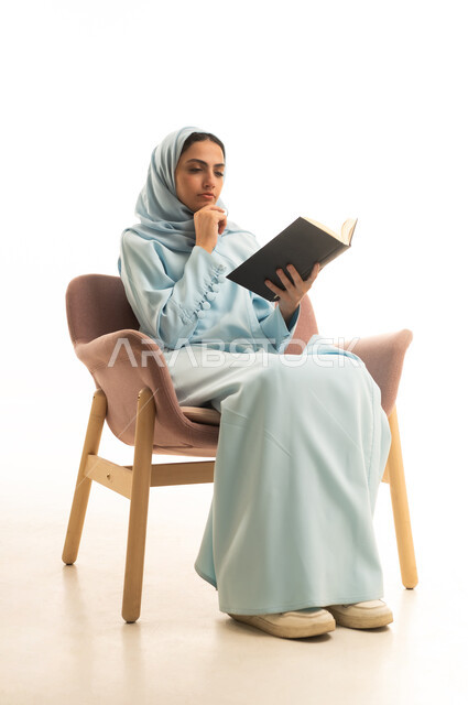 قراءة الروايات والقصص الممتعة،  قضاء وقت الفراغ بالتعلم والثقافة، القراءة بتمعن وتركيز، بورتريه لإمراة عربية سعودية خليجية محجبة ترتدي العباءة تجلس على كرسي تحمل بيدها كتاب تثقيفي، خلفية بيضاء