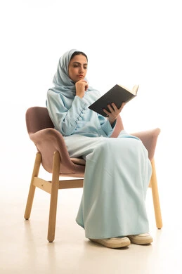 قراءة الروايات والقصص الممتعة،  قضاء وقت الفراغ بالتعلم والثقافة، القراءة بتمعن وتركيز، بورتريه لإمراة عربية سعودية خليجية محجبة ترتدي العباءة تجلس على كرسي تحمل بيدها كتاب تثقيفي، خلفية بيضاء