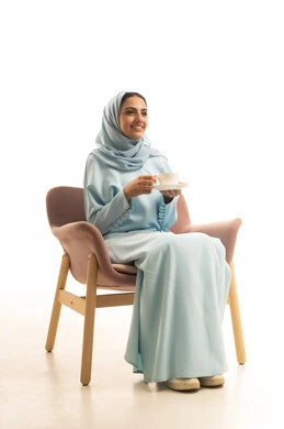 Taste the distinctive flavor of the hot drink, mood adjustment by drinking caffeine, a portrait of a Saudi Gulf Arab woman wearing a hijab and an abaya sitting on a chair holding a cup of coffee, gestures of happiness and joy, white background.