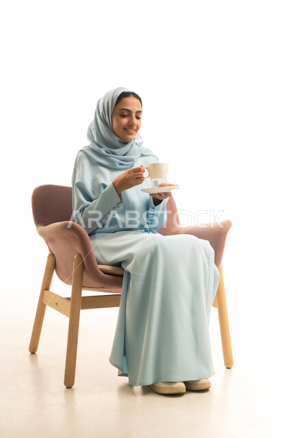 الاستمتاع بتناول المشروبات الساخنة، تعديل المزاج بشرب الكافيين، بورتريه لإمرأة عربية خليجية سعودية محجبة ترتدي العباءة تجلس على كرسي تحمل كوب من القهوة، ايماءات السعادة والسرور، خلفية بيضاء