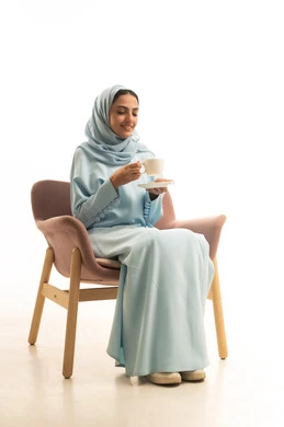Enjoying hot beverages, mood enhancement by drinking caffeine, a portrait of a Saudi Arabian Gulf woman wearing a hijab and an abaya sitting on a chair holding a cup of coffee, gestures of happiness and joy, white background.