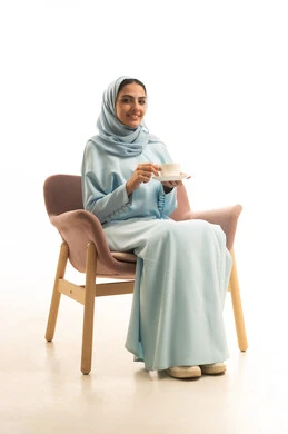 Mood adjustment by drinking caffeine, enjoying hot beverages, a portrait of a Saudi Arabian Gulf woman wearing a hijab and an abaya sitting on a chair holding a cup of coffee, gestures of happiness and joy, white background.