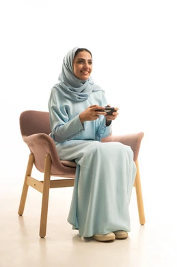 إيماءات وتعابير وجه ويدين تدل الاندماج و التركيز، قضاء أوقات الفراغ في الفيديو جيمز، بورتريه لسيده عربيه سعودية خليجية ترتدي الحجاب والعباءة تجلس على كرسي تحمل بيدها عصا التحكم وتلعب بألعاب الفيديو، خلفية بيضاء
