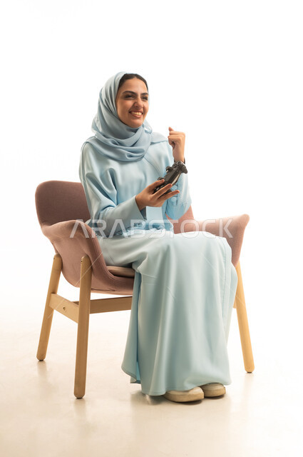 قضاء أوقات الفراغ في الفيديو جيمز، إيماءات وتعابير وجه ويدين تدل على الفوز والانتصار، بورتريه لسيده عربيه سعودية خليجية ترتدي الحجاب والعباءة تجلس على كرسي تحمل بيدها عصا التحكم وتلعب بألعاب الفيديو، خلفية بيضاء