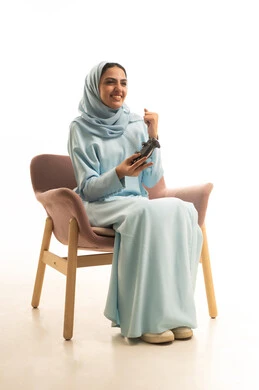 قضاء أوقات الفراغ في الفيديو جيمز، إيماءات وتعابير وجه ويدين تدل على الفوز والانتصار، بورتريه لسيده عربيه سعودية خليجية ترتدي الحجاب والعباءة تجلس على كرسي تحمل بيدها عصا التحكم وتلعب بألعاب الفيديو، خلفية بيضاء