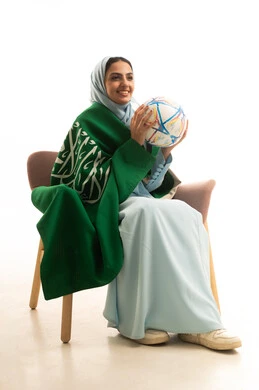ذكرى يوم العلم 11 مارس، اليوم الوطني السعودي 23 سبتمبر،  النظر بإيماءات البهجة والفرح بالانتصارفي مباراة كرة القدم، بورتريه لامراه عربيه خليجيه سعودية محجبة ترتدي العباءة تجلس على كرسي  تحمل كره القدم، خلفية بيضاء