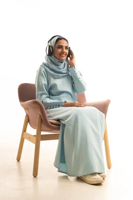 Enjoying listening to favorite songs, using modern headphone technology, expressions of immersion and interaction with musical tunes, a portrait of a Gulf Arab woman wearing a hijab and an abaya sitting on a chair with headphones on, white background.