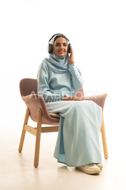 Using modern headphone technology, expressions of immersion and interaction with musical tunes, enjoying listening to favorite songs, a portrait of a Gulf Arab woman wearing a hijab and an abaya sitting on a chair with headphones on, white background.
