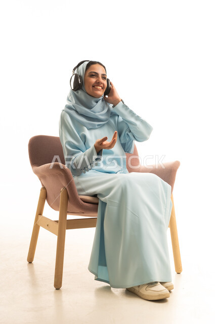تعابير الاندماج و التفاعل مع الأنغام الموسيقية، استخدام تقنية الهيدفون الحديثة, الاستمتاع بسماع الأغاني المفضلة, بورتريه لامرأة عربية خليجية محجبة ترتدي العباءة تجلس على كرسي تضع سماعات الرأس الصوتية، خلفية بيضاء