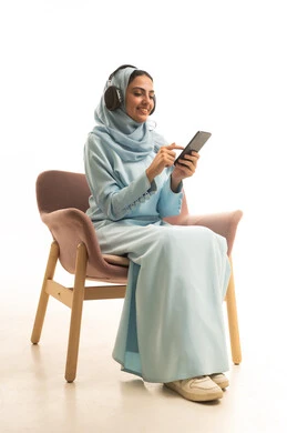 استخدام جهاز تقني حديث، الاستماع الى الاغاني والموسيقى، بورتريه لإمرأة عربية خليجية سعودية ترتدي الحجاب والعباءة تجلس على كرسي تضع سماعات الهيدفون تمسك بيدها الهاتف المحمول، تعابير السرور والسعادة، خلفية بيضاء