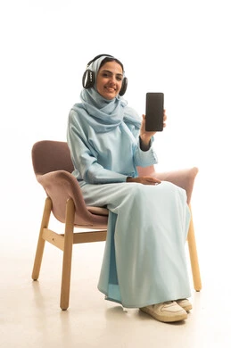 Looking at the camera and displaying a blank black screen through the mobile phone, using advanced modern technology and devices, a portrait of a smiling Saudi Arabian Gulf woman wearing a hijab, dressed in an abaya and headphones, sitting on a chair, with a white background.